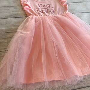 May include: A pink tulle dress with a pink sleeveless top featuring the text "Big Sister" in pink glitter.