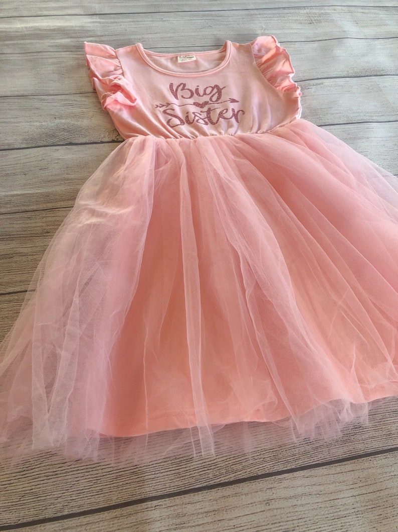 May include: A pink tulle dress with a pink sleeveless top featuring the text "Big Sister" in pink glitter.