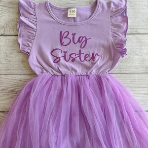 May include: A purple tulle tutu dress with a lavender short-sleeved shirt that says "Big Sister" in sparkly pink letters.