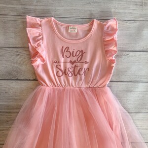 May include: A pink tulle dress with a pink short-sleeved shirt top. The shirt has a glittery pink design that says "Big Sister" with an arrow and a heart.