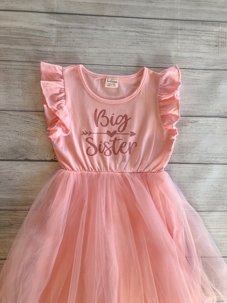 May include: A pink tulle dress with a pink short-sleeved shirt top. The shirt has a glittery pink design that says "Big Sister" with an arrow and a heart.