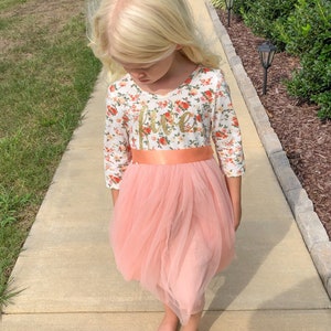 May include: A young girl wearing a white dress with a floral pattern and a pink tulle skirt. The dress has a peach-colored sash and the word "five" written in gold glitter on the front.