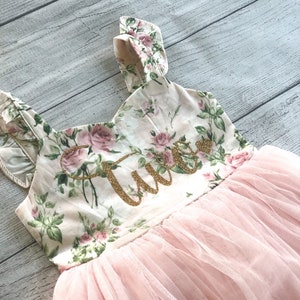 Op de afbeelding: Een roze tulejurk met bloemenprint en een gouden glitter "Twee" op het lijfje.