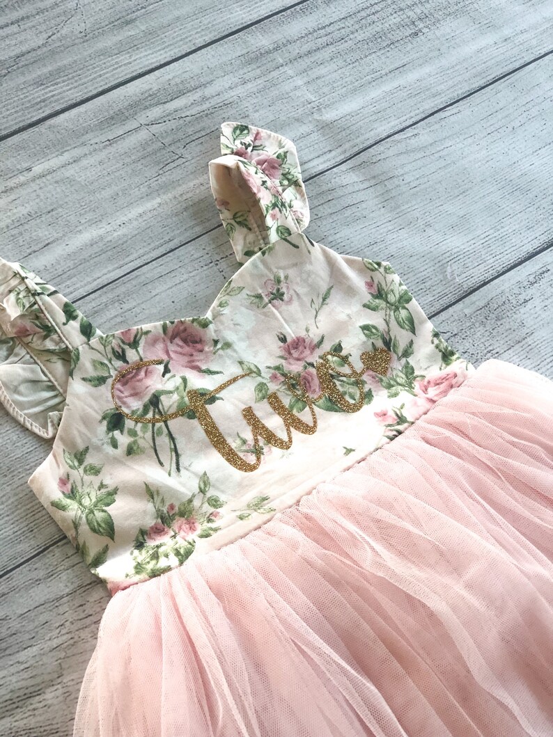Op de afbeelding: Een roze tulejurk met bloemenprint en een gouden glitter "Twee" op het lijfje.