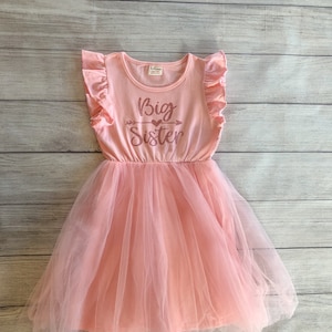 May include: A pink tulle dress with a pink short-sleeved shirt overlay. The shirt has a ruffled neckline and the text "Big Sister" with a heart and arrow graphic printed on the front.