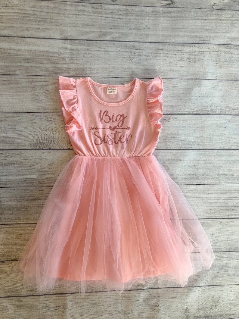 May include: A pink tulle dress with a pink short-sleeved shirt overlay. The shirt has a ruffled neckline and the text "Big Sister" with a heart and arrow graphic printed on the front.