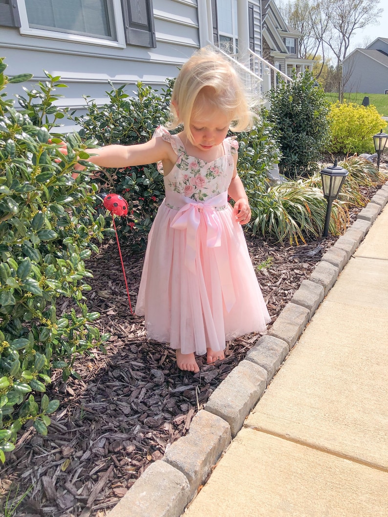 Op de afbeelding: Een jong meisje draagt een roze tulejurk met een bloemenprint en een roze lint om de taille. Ze staat op een pad met bruine mulch en een groene struik achter haar.