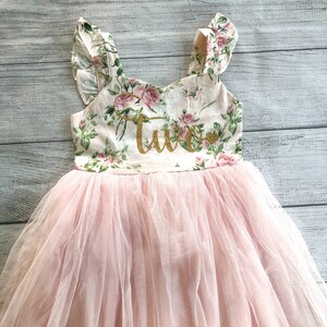 Op de afbeelding: Een roze tule tutu rok met een bloemetjesprint bovenstuk en het woord "Twee" in gouden glitter. Het bovenstuk heeft gerimpelde bandjes.