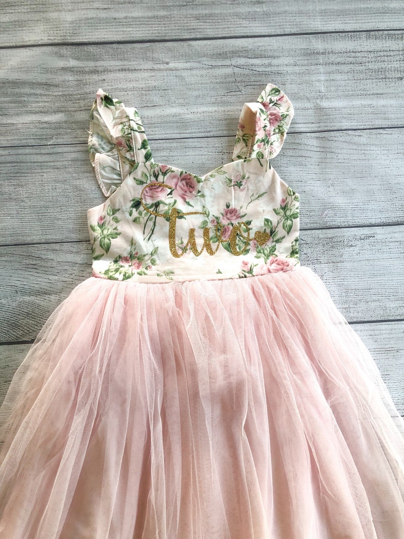 Op de afbeelding: Een roze tule tutu rok met een bloemetjesprint bovenstuk en het woord "Twee" in gouden glitter. Het bovenstuk heeft gerimpelde bandjes.