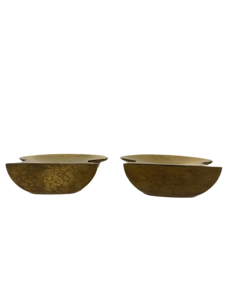 May include: Two brass-colored, semi-circular decorative bowls. Each bowl has a textured surface and a flat, horizontal top. The bowls are positioned side-by-side against a white background.