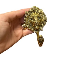May include: A brass lion head hook with a detailed mane and a hook below. The hook is a golden color and is likely used for hanging items. The lion's face is intricately designed, giving it a regal appearance.