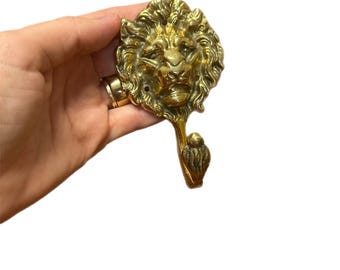May include: A brass lion head hook with a detailed mane and a hook below. The hook is a golden color and is likely used for hanging items. The lion's face is intricately designed, giving it a regal appearance.
