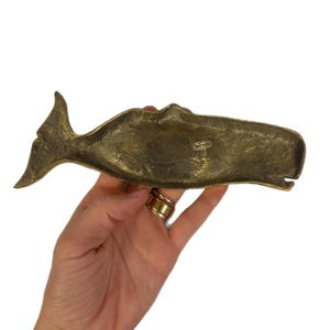 K&ouml;nnte beinhalten: Eine Messingwal-Figur mit strukturierter Oberfl&auml;che. Die Wal ist horizontal positioniert, wobei Schwanz und Kopf sichtbar sind. Die Figur hat ein Vintage-Aussehen und wird in einer Hand vor wei&szlig;em Hintergrund gehalten.