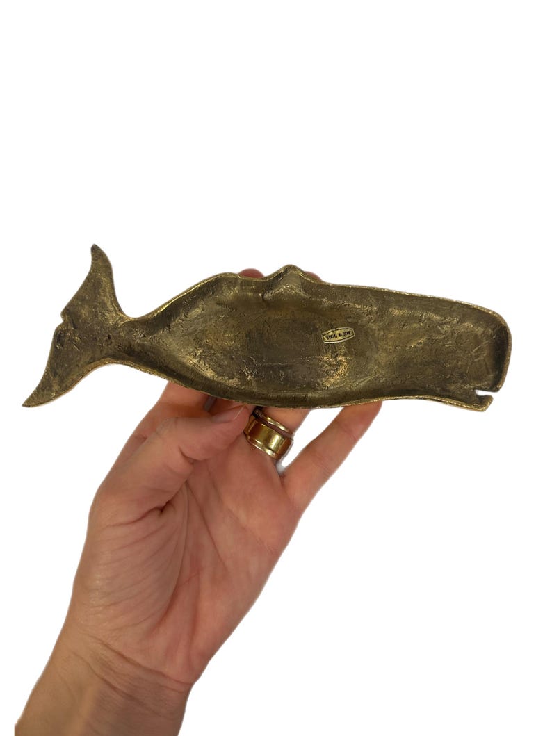 K&ouml;nnte beinhalten: Eine Messingwal-Figur mit strukturierter Oberfl&auml;che. Die Wal ist horizontal positioniert, wobei Schwanz und Kopf sichtbar sind. Die Figur hat ein Vintage-Aussehen und wird in einer Hand vor wei&szlig;em Hintergrund gehalten.