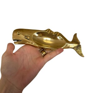 K&ouml;nnte beinhalten: Ein goldfarbener Aschenbecher in Walform mit einer breiten, offenen Schale. Der Wal hat detaillierte Flossen und eine strukturierte Schwanzflosse. Der Aschenbecher wird in einer Hand vor wei&szlig;em Hintergrund gehalten. Das Maul des Wals ist ge&ouml;ffnet.