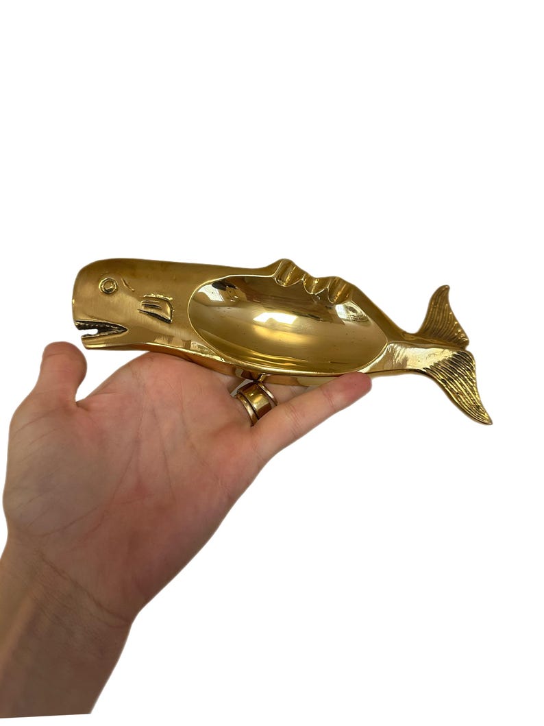 K&ouml;nnte beinhalten: Ein goldfarbener Aschenbecher in Walform mit einer breiten, offenen Schale. Der Wal hat detaillierte Flossen und eine strukturierte Schwanzflosse. Der Aschenbecher wird in einer Hand vor wei&szlig;em Hintergrund gehalten. Das Maul des Wals ist ge&ouml;ffnet.