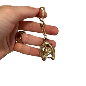 May include: A gold-colored keychain featuring a horseshoe design with a horse head in the center. The keychain has a clasp for easy attachment. The design is detailed and the metal has a polished finish. The keychain is held by a hand.