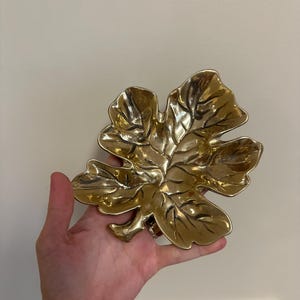 以下が含まれることがあります： 金色の葉の形をした装飾的な皿。 皿には詳細な葉脈があり、わずかに凹んだ形をしています。 金属仕上げはヴィンテージまたはアンティークの外観を与えます。 皿は手で持たれています。
