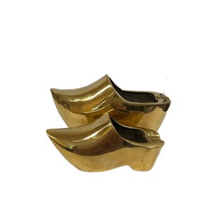 May include: A pair of shiny, golden-colored clogs, a traditional Dutch wooden shoe. The clogs are made of a solid material and have a smooth, reflective surface. The shoes are positioned side by side.