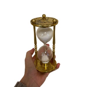 May include: A gold-coloured hourglass with white sand inside. The hourglass is held in a hand and has a decorative top and base with Roman numerals. The sand is flowing from the top to the bottom chamber.