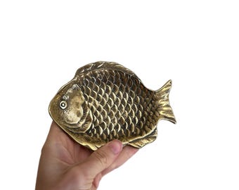 May include: A brass fish-shaped dish featuring a detailed scale pattern across its body, a defined head with a circular eye, and a textured fin design. The metallic surface has a polished finish with natural variations in tone and luster.