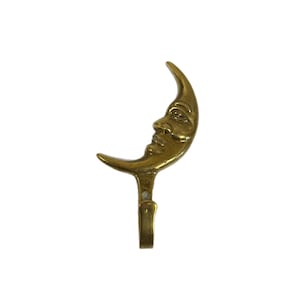 Puede incluir: Un colgador de pared de color latón con forma de media luna con rostro humano. Este colgador decorativo presenta un rostro detallado en la superficie de la luna y un pequeño gancho en la parte inferior para colgar objetos. El colgador mide unos 10 cm de largo.