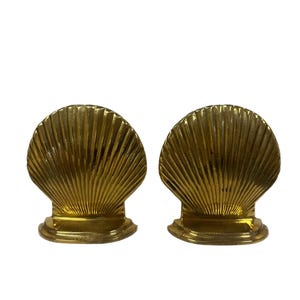 May include: Two gold-colored seashell-shaped bookends. Each bookend has a fan-like design with vertical ridges and a small, rounded base. The bookends are made of a shiny, metallic material.