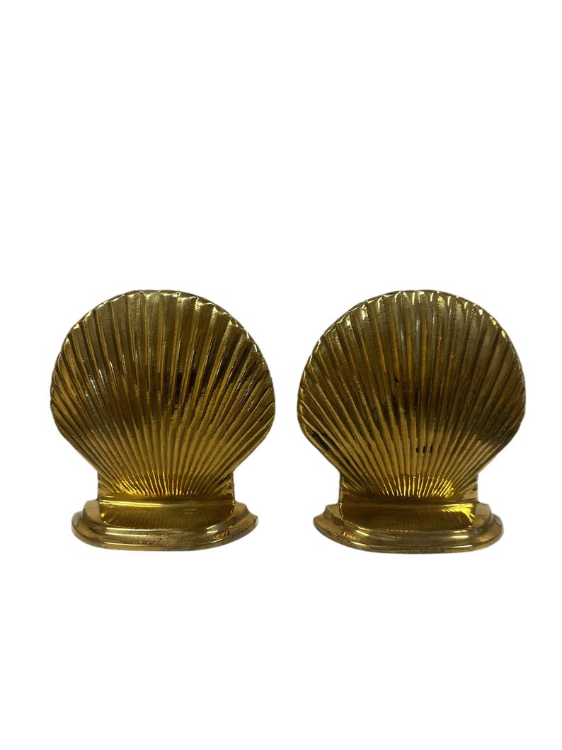 May include: Two gold-colored seashell-shaped bookends. Each bookend has a fan-like design with vertical ridges and a small, rounded base. The bookends are made of a shiny, metallic material.