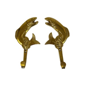 May include: Two brass-colored wall hooks, each shaped like a fish leaping upwards. The hooks have a detailed design, with visible scales and fins. The hooks are mounted on a white background.
