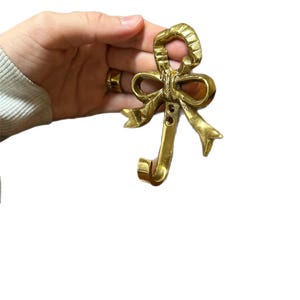 Pode incluir: Um gancho em forma de laço de cor latão. O gancho tem a forma de um laço com uma argola na parte superior e um gancho curvo na parte inferior. O laço tem dobras de fita detalhadas e três pequenos orifícios.