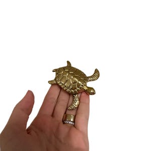 Könnte beinhalten: Eine goldfarbene Schildkrötenfigur mit detailliertem Panzer und Flossen. Die Schildkröte wird in einer Hand gehalten, und zwei Ringe sind an einem Finger zu sehen. Die Ringe sind goldfarben, einer mit einem Band aus kleinen, klaren Steinen.