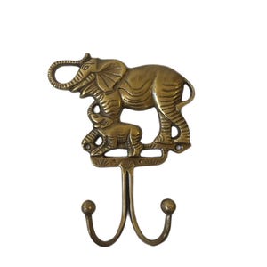 May include: A brass-colored wall hook featuring an elephant and baby elephant design. The hook has two curved prongs with rounded tips, suitable for hanging items. The elephant figures are detailed with etched lines.