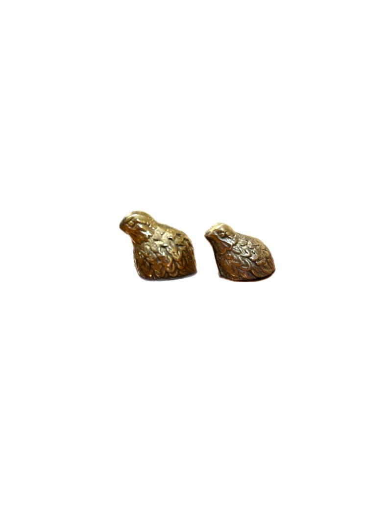 May include: Two small, detailed brass quail figurines. The birds are in a resting position, with textured feathers and a warm, metallic sheen. They are set against a plain white background, highlighting their intricate design.