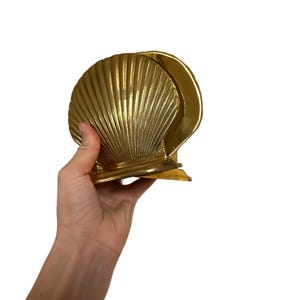 May include: A gold-colored seashell-shaped napkin holder. The holder is made of metal and has a ribbed texture. The design is a stylized scallop shell, with a curved back and a flat base. The holder is held by a hand.