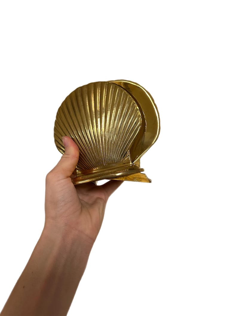 May include: A gold-colored seashell-shaped napkin holder. The holder is made of metal and has a ribbed texture. The design is a stylized scallop shell, with a curved back and a flat base. The holder is held by a hand.
