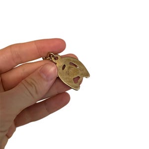 May include: A gold-colored metal pendant in the shape of a stylized face, held by a hand against a white background. The pendant has cut-out details for the eyes and mouth, and a small loop at the top for a chain.