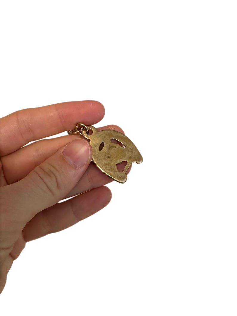 May include: A gold-colored metal pendant in the shape of a stylized face, held by a hand against a white background. The pendant has cut-out details for the eyes and mouth, and a small loop at the top for a chain.