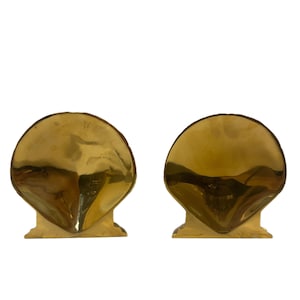 May include: Two gold-colored seashell bookends. Each bookend has a rounded, fan-shaped top and a rectangular base. The bookends are made of a shiny, reflective material and are set against a white background.