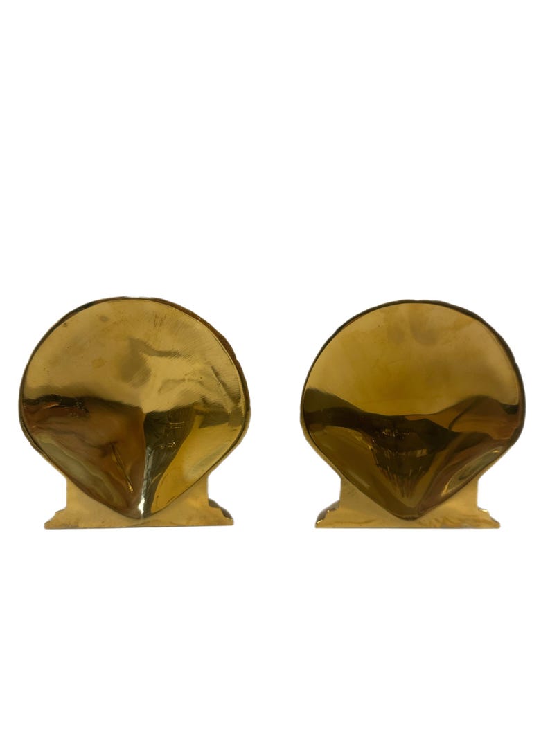 May include: Two gold-colored seashell bookends. Each bookend has a rounded, fan-shaped top and a rectangular base. The bookends are made of a shiny, reflective material and are set against a white background.