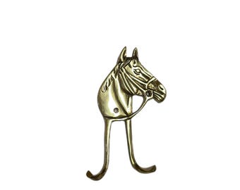 May include: A brass-colored wall hook shaped like a horse's head with a bridle. The hook extends downward with two curved arms. The horse's head is detailed with etched lines, and the hook is mounted on a white background.