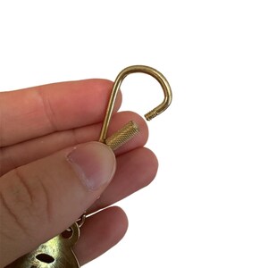May include: A gold-colored keychain hook with a textured screw-on base. The hook is open, revealing the threaded end. The keychain has a decorative element at the bottom, with a cutout design.