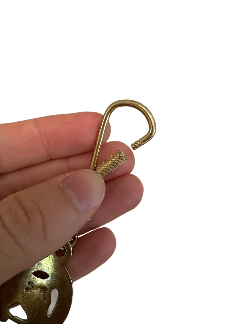 May include: A gold-colored keychain hook with a textured screw-on base. The hook is open, revealing the threaded end. The keychain has a decorative element at the bottom, with a cutout design.