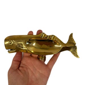 K&ouml;nnte beinhalten: Ein messingfarbener Aschenbecher in Walform. Der Aschenbecher hat eine gl&auml;nzende goldene Farbe und zeigt ein detailliertes Design eines Wals mit breitem Maul und fischflossenf&ouml;rmigem Schwanz. Der Aschenbecher wird in einer Hand gehalten.