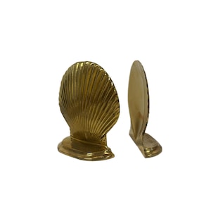 Puede incluir: Dos sujetalibros con forma de concha marina de latón. Los sujetalibros son de color dorado y tienen una textura acanalada detallada. Están diseñados para colocarse en posición vertical y añadir un toque decorativo a cualquier estantería o escritorio.