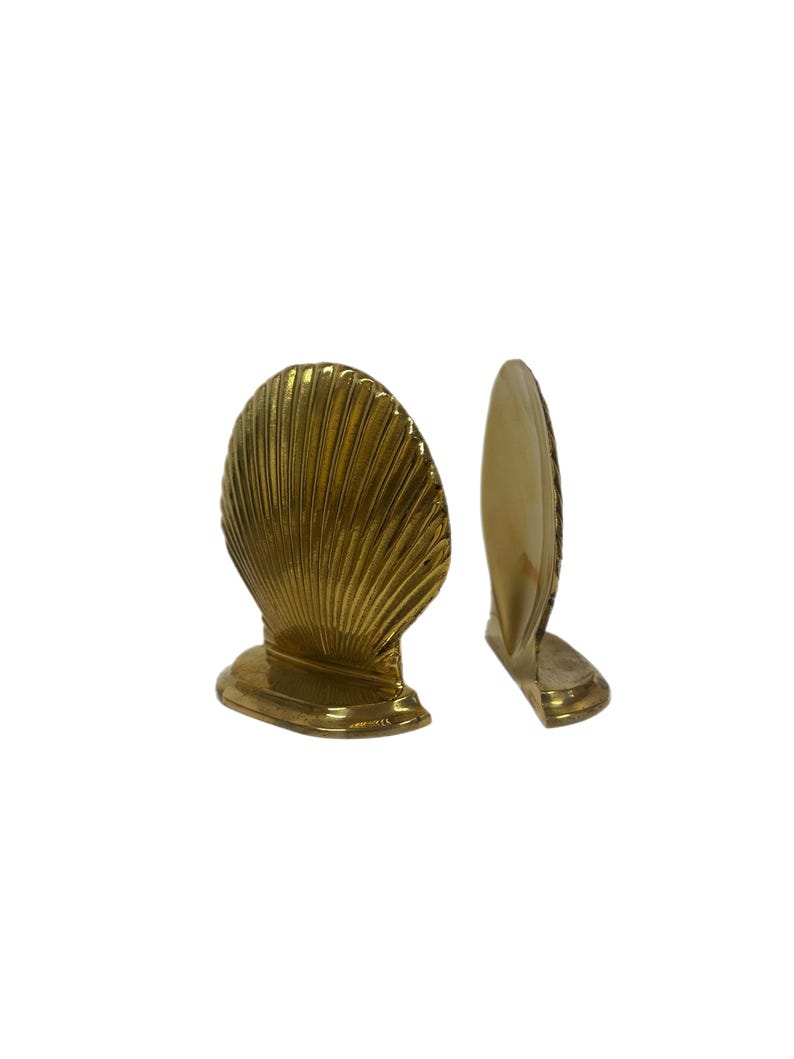 May include: Two brass seashell-shaped bookends. The bookends are golden in color and have a detailed, ribbed texture. They are designed to stand upright and add a decorative touch to any bookshelf or desk.