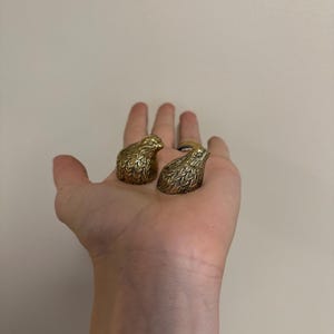 May include: Two brass bird-shaped rings, each with detailed feather textures, are displayed. The rings are held in a hand against a neutral background. The rings are likely for jewelry.