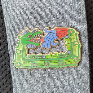 May include: A gold-colored pin with a green and blue design depicting a mythical creature with a feathered headdress. The pin is attached to a gray fabric strap.