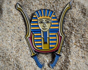 Puede incluir: Pin de esmalte dorado y azul de un faraón egipcio con un tocado tradicional. El pin tiene forma de escarabajo con dos espadas a cada lado.