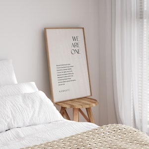 May include: A framed print with a white background and black text that reads "We Are One." The text is a poem about honoring the place in your soul where the entire universe resides. The print is hanging on a white wall above a wooden stool.