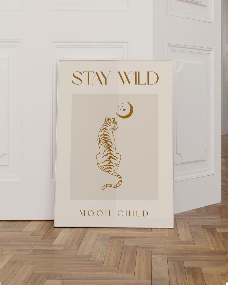 May include: A beige poster with the text "STAY WILD" in brown lettering at the top. A brown outline of a tiger sitting facing away from the viewer is in the center of the poster. A crescent moon and stars are above the tiger. The text "MOON CHILD" is at the bottom of the poster in brown lettering.